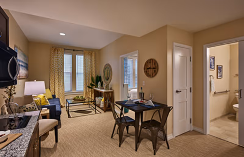 Furnished apartment-style living area with a small dining table, sofa and kitchenette, and a doorway leading to a bathroom.