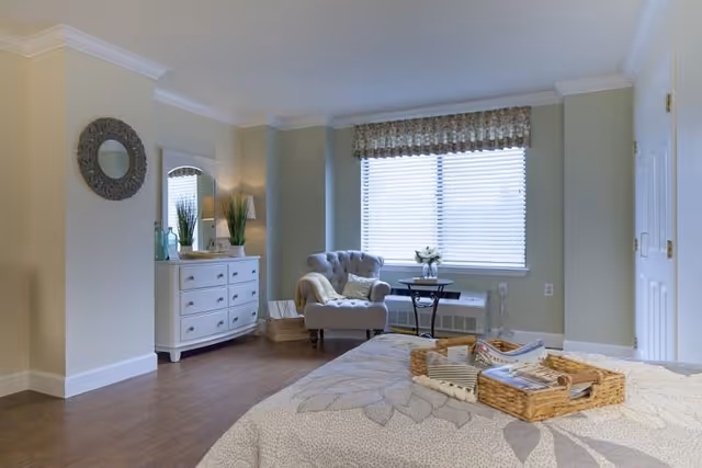 Sunny, cozy bedroom with a bed in the foreground, a tufted armchair by the window, a white dresser, and hardwood floors.