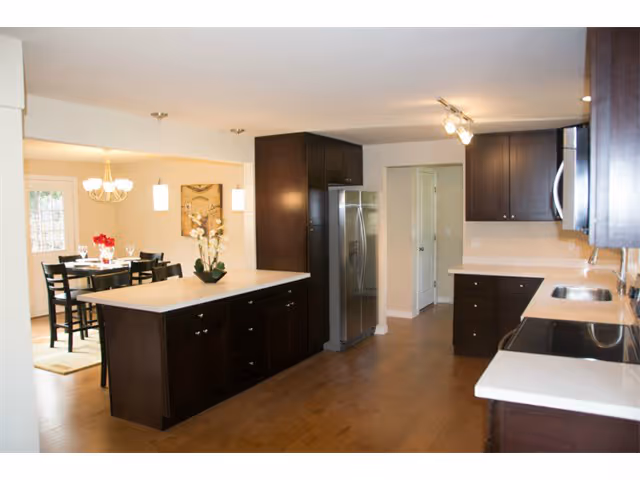 A modern kitchen with dark wood cabinets, white countertops, and stainless steel appliances including a refrigerator and stove. The kitchen opens into a dining area with a table set for six, featuring a chandelier and pendant lights. The floor is wooden and the walls are painted light beige.