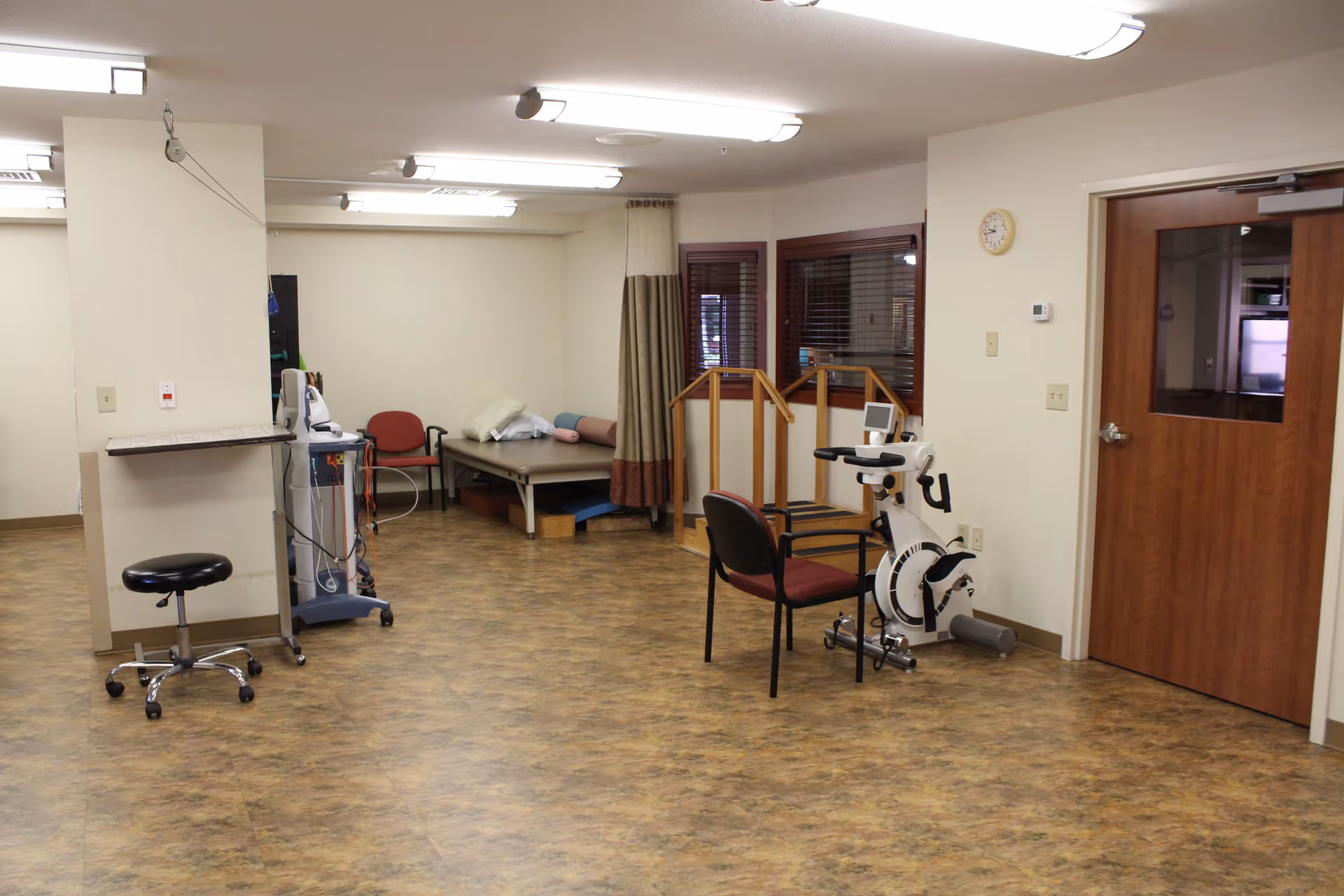 A physical therapy or rehabilitation room with exercise equipment including a stationary bike, parallel bars for walking practice, a treatment table with pillows, a rolling stool, and a chair. The room has wood-patterned flooring, beige walls, fluorescent ceiling lights, and a wooden door with a window.