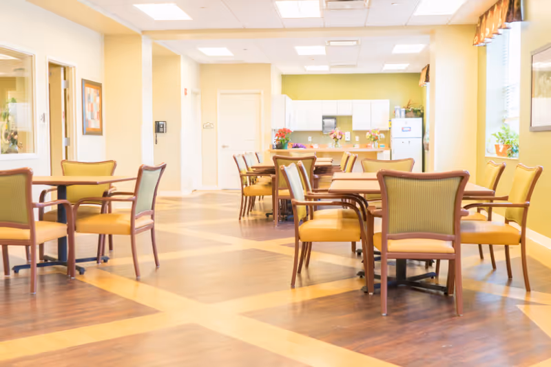 Bright communal dining area with multiple tables and chairs and a kitchenette in the background.