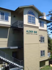 Exterior view of a multi-story building with beige siding and multiple windows, featuring a sign that reads 'Alder Bay Assisted Living'. There are metal staircases and railings visible on the left side of the building, with trees and a clear blue sky in the background.