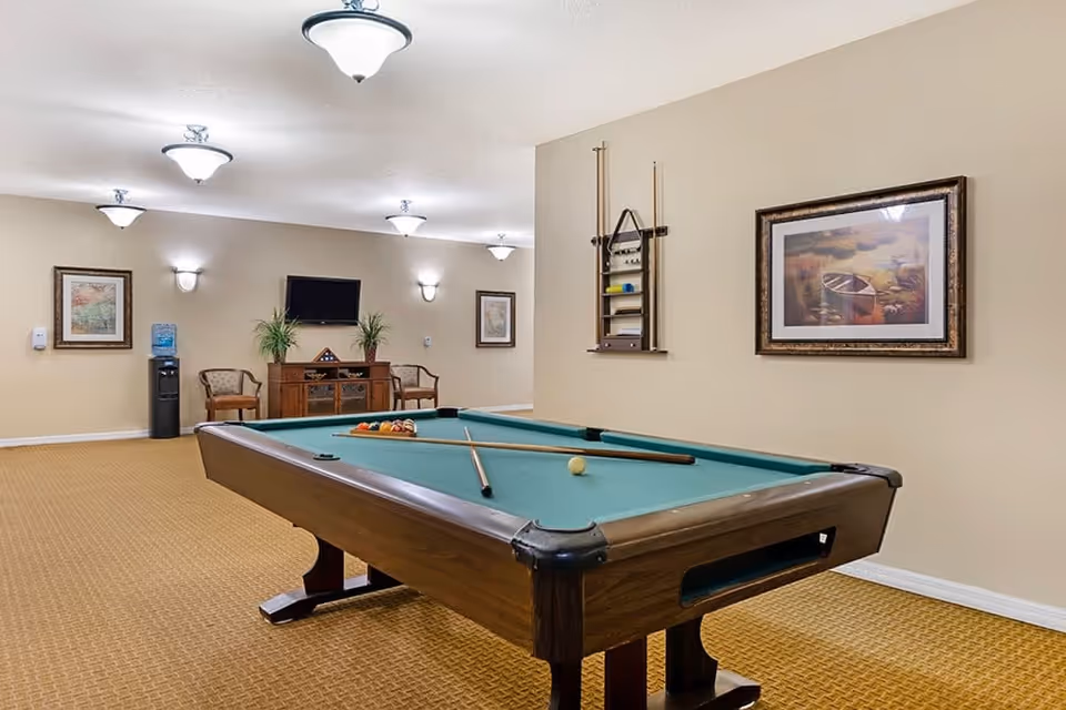 A well-lit recreational room featuring a pool table with cues and balls arranged on it. The room has beige walls adorned with framed artwork and a mounted pool cue rack. In the background, there is a wooden cabinet with a flat-screen TV above it, two chairs, a water dispenser, and additional framed pictures on the walls.