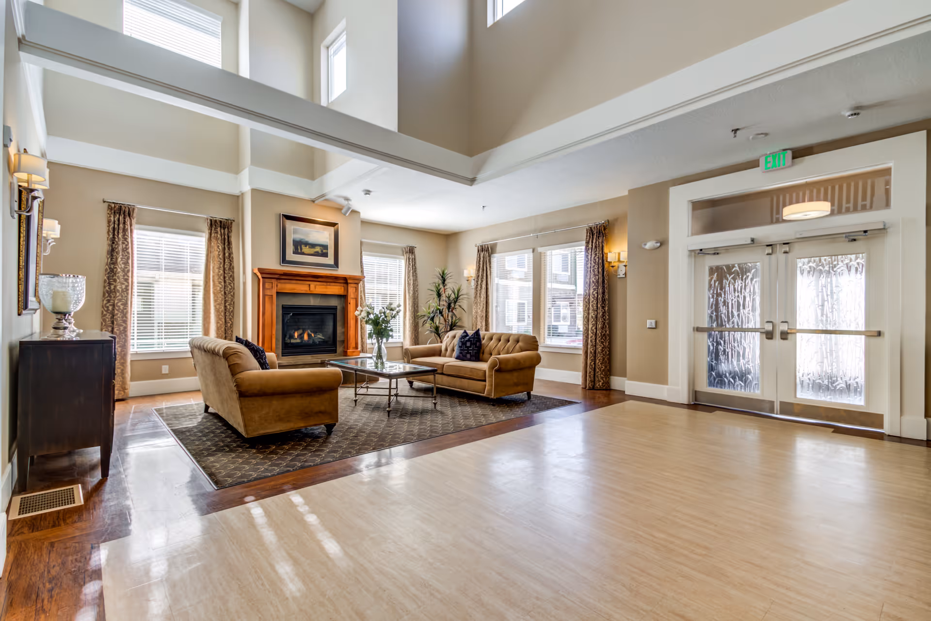 A spacious senior living facility common area with high ceilings and large windows allowing natural light. The room features a cozy seating area with two beige sofas, a glass coffee table, a fireplace with a wooden mantel, and decorative plants. There are double glass doors with frosted patterns on the right side and beige walls with curtains on the windows.