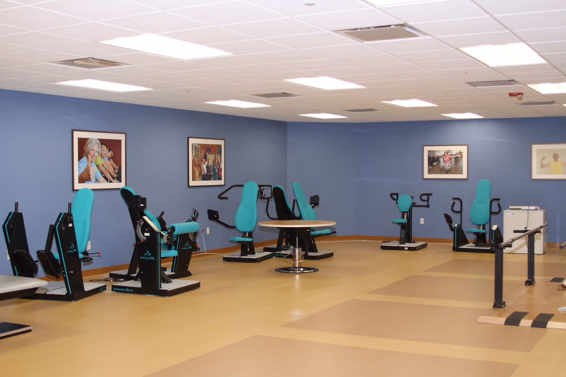 Rehabilitation exercise room with teal exercise machines, parallel bars, and framed photos on blue walls.