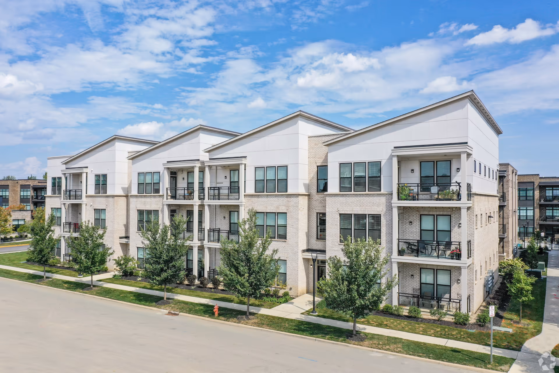 Three-story modern apartment building with balconies, landscaping and trees along the sidewalk under a blue sky.