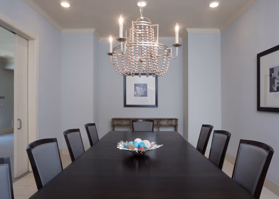 Long dark dining table with chairs and a decorative bowl beneath a beaded chandelier in a neutral-toned dining room.