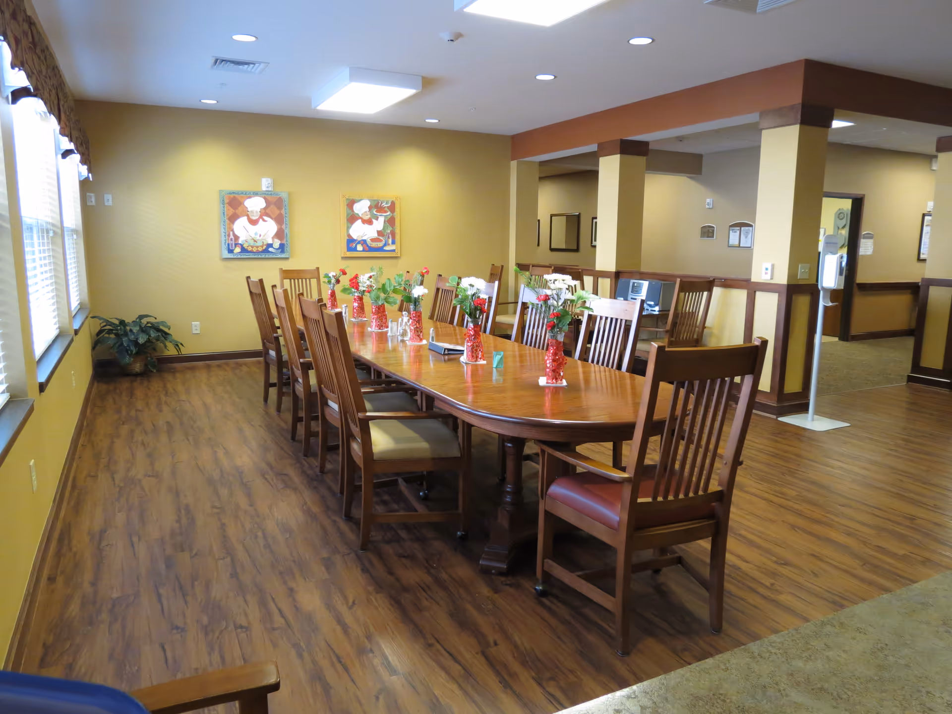 A dining room with a long wooden table surrounded by wooden chairs with cushions. The table is decorated with small vases of flowers. The room has wood flooring, yellow walls, and two framed pictures of chefs on the far wall. Large windows with blinds allow natural light to enter the space.