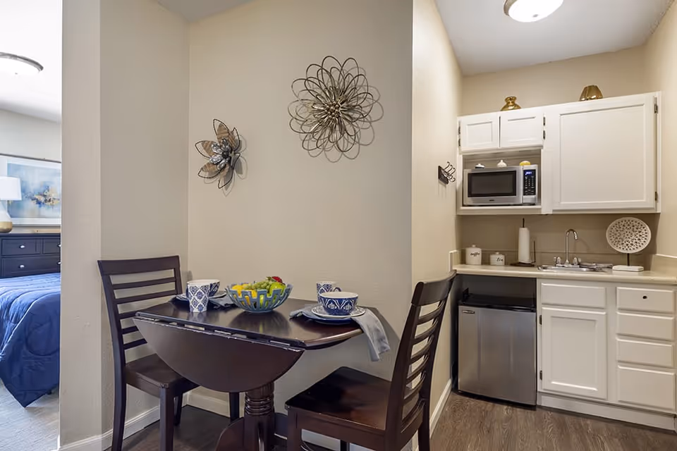 Small kitchenette and dining area with a round wooden table set for two, two chairs, and a compact kitchen with white cabinets, microwave, and mini fridge.