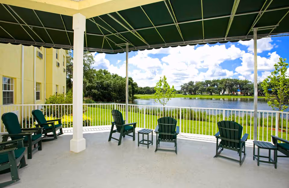 Covered outdoor patio area with green Adirondack chairs and small tables overlooking a lake with trees and a bright blue sky with clouds.