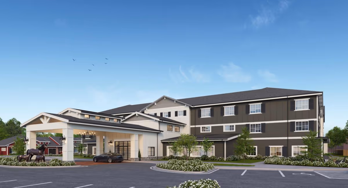 Exterior view of The Inn at University Village Assisted Living Community building with a covered entrance, landscaped greenery, and a parking lot under a clear blue sky.