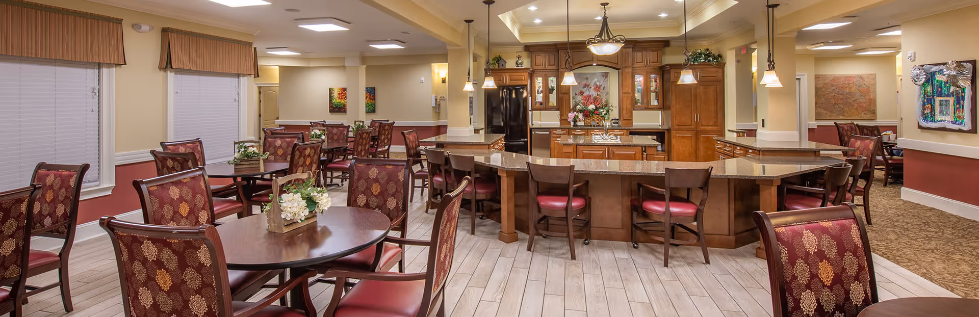 Spacious senior living dining area with round tables and patterned chairs surrounding a central kitchen island and warm lighting.
