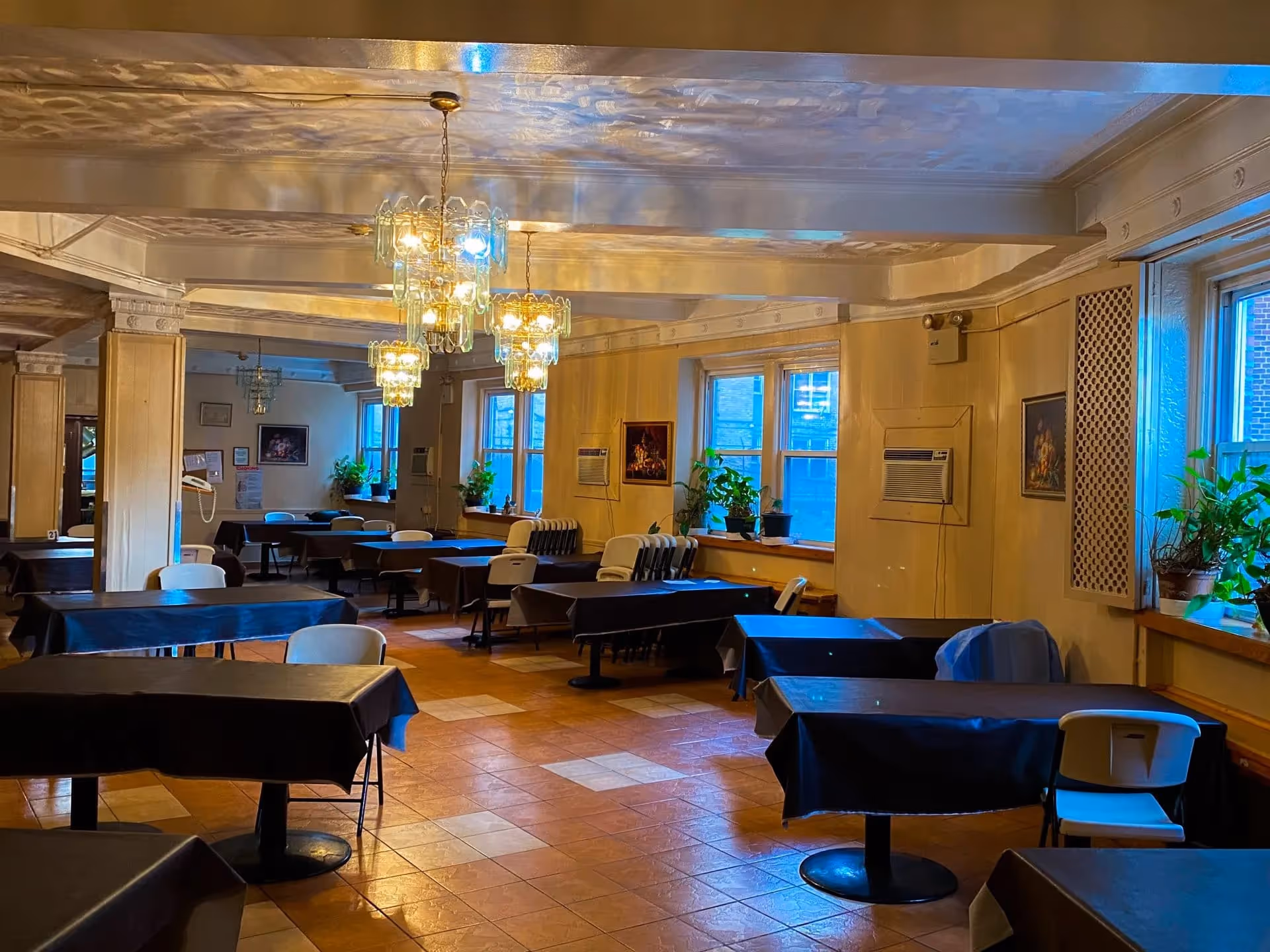 Interior view of a dining room with multiple tables covered in dark tablecloths and white chairs. The room has large windows with potted plants on the windowsills, beige walls, and decorative chandeliers hanging from the ceiling. There are framed pictures on the walls and an air conditioning unit installed in one window.