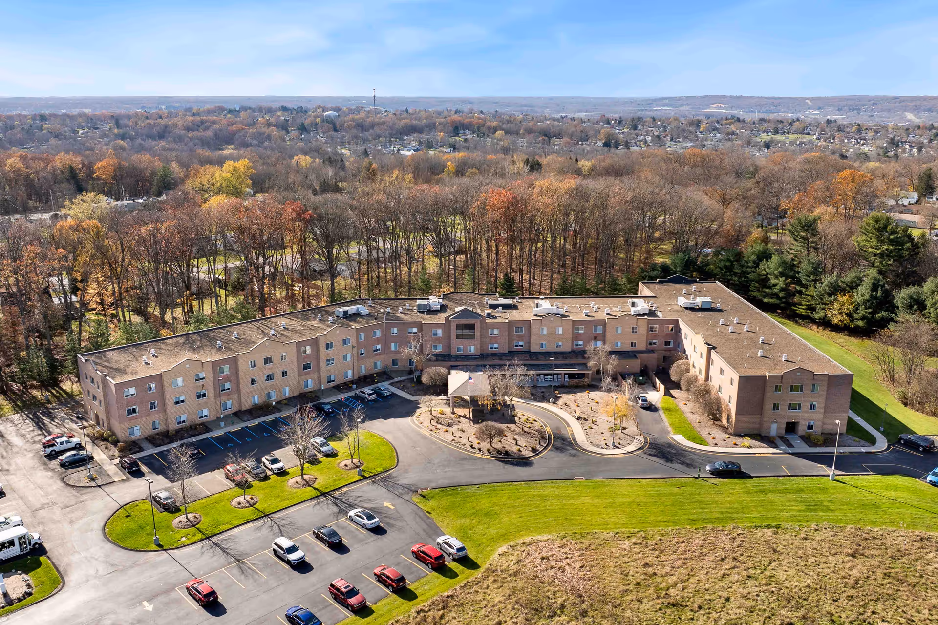 Aerial view of a large, three-story senior living facility surrounded by trees with autumn foliage. The building is shaped like a wide U with a parking lot in front, several cars parked, and landscaped areas with small trees and bushes. The background shows a suburban area with more trees and houses extending to the horizon.