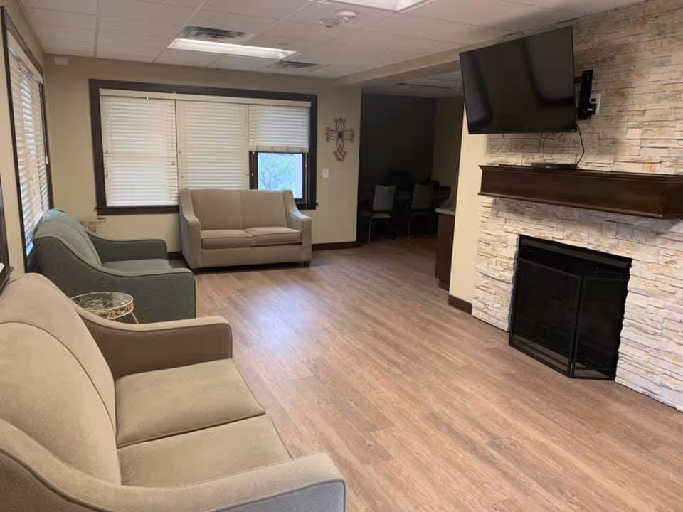 A cozy living room with three sofas arranged around a stone fireplace with a mounted flat-screen TV above it. The room has wood flooring, beige walls, and windows with blinds letting in natural light. There is a small side table next to one of the sofas and a decorative wall piece near the entrance to another room.