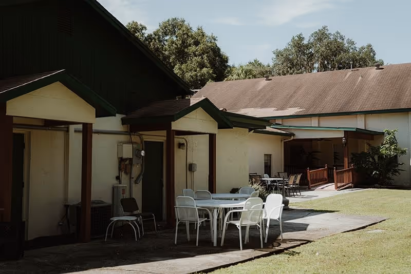 Outdoor patio area at Prestige Manor Assisted Living Facility Belleview with white chairs and tables on a concrete surface next to a beige building with green trim, surrounded by grass and trees under a clear sky.