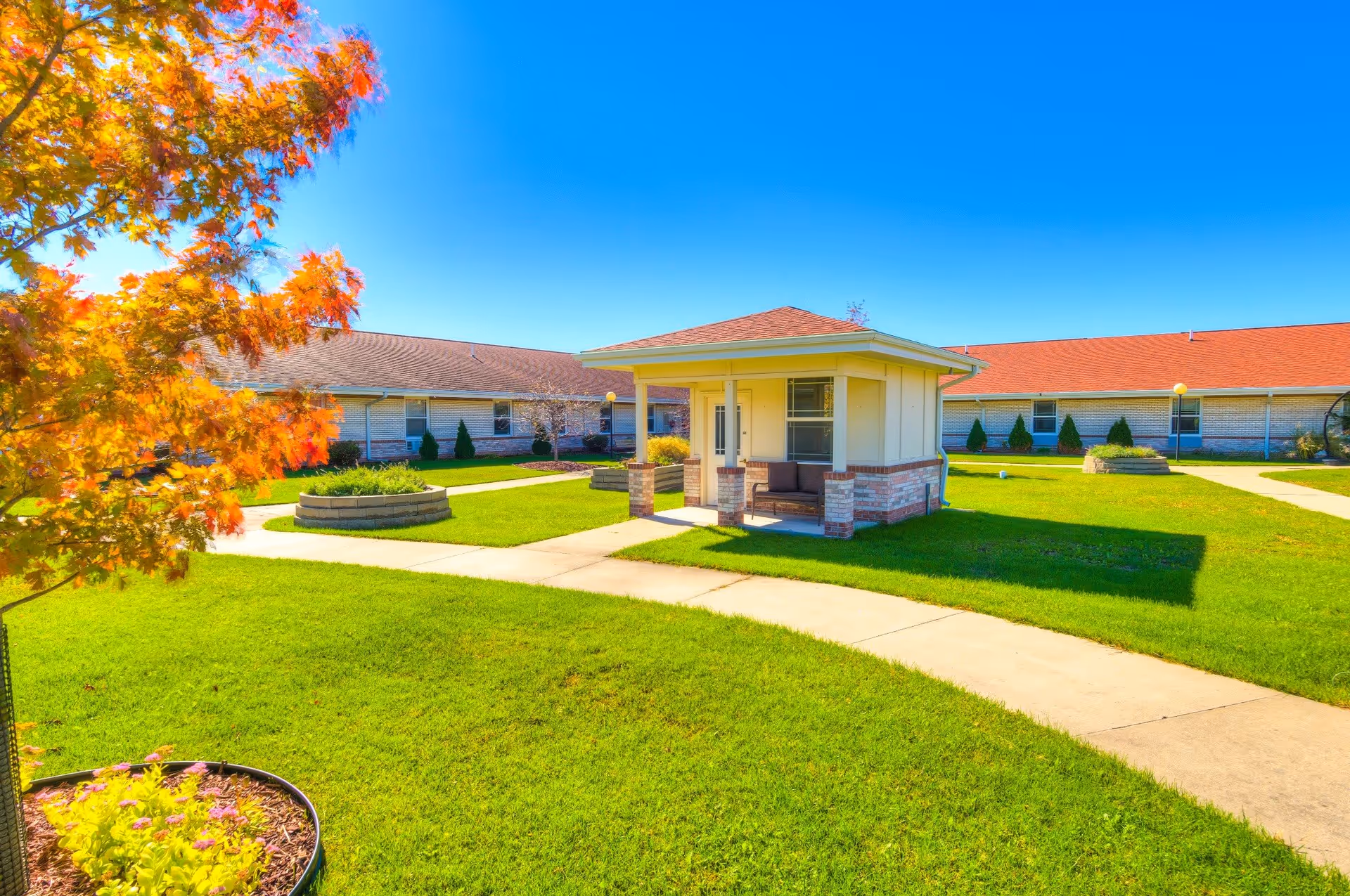 A bright outdoor courtyard area with green grass, a small covered seating structure with a bench, paved walkways, and a tree with autumn-colored leaves. The courtyard is surrounded by single-story buildings with red roofs under a clear blue sky.