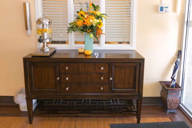 A dark wooden console table in an entry area with a vase of flowers, a lemon water dispenser, small decorative pumpkins, and an umbrella stand by the door.
