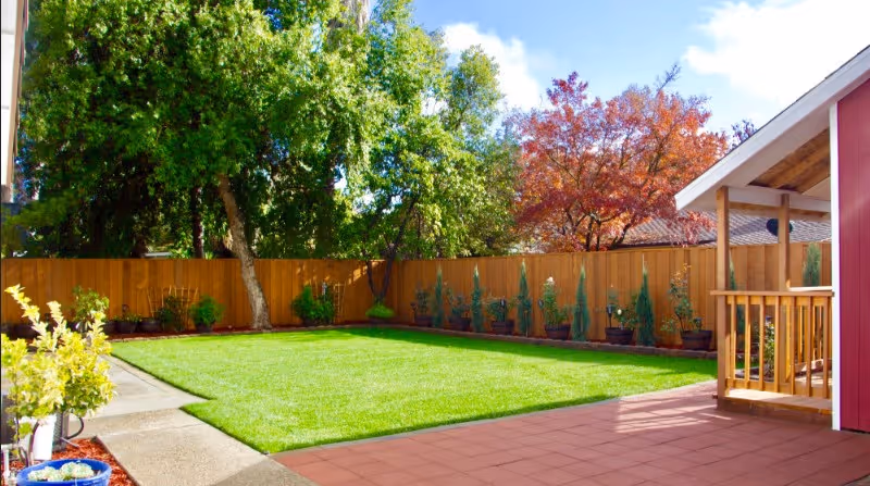 A sunny outdoor area featuring a well-maintained green lawn bordered by a wooden fence. There are various potted plants along the fence and large trees providing shade. On the right side, there is a red building with a small porch and wooden railing. The sky is blue with some clouds.