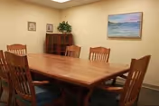 A small meeting or dining room with a wooden rectangular table surrounded by six wooden chairs with blue cushions. There is a wooden cabinet with glass doors in the corner, a potted plant on top, and a framed landscape painting on the beige wall.