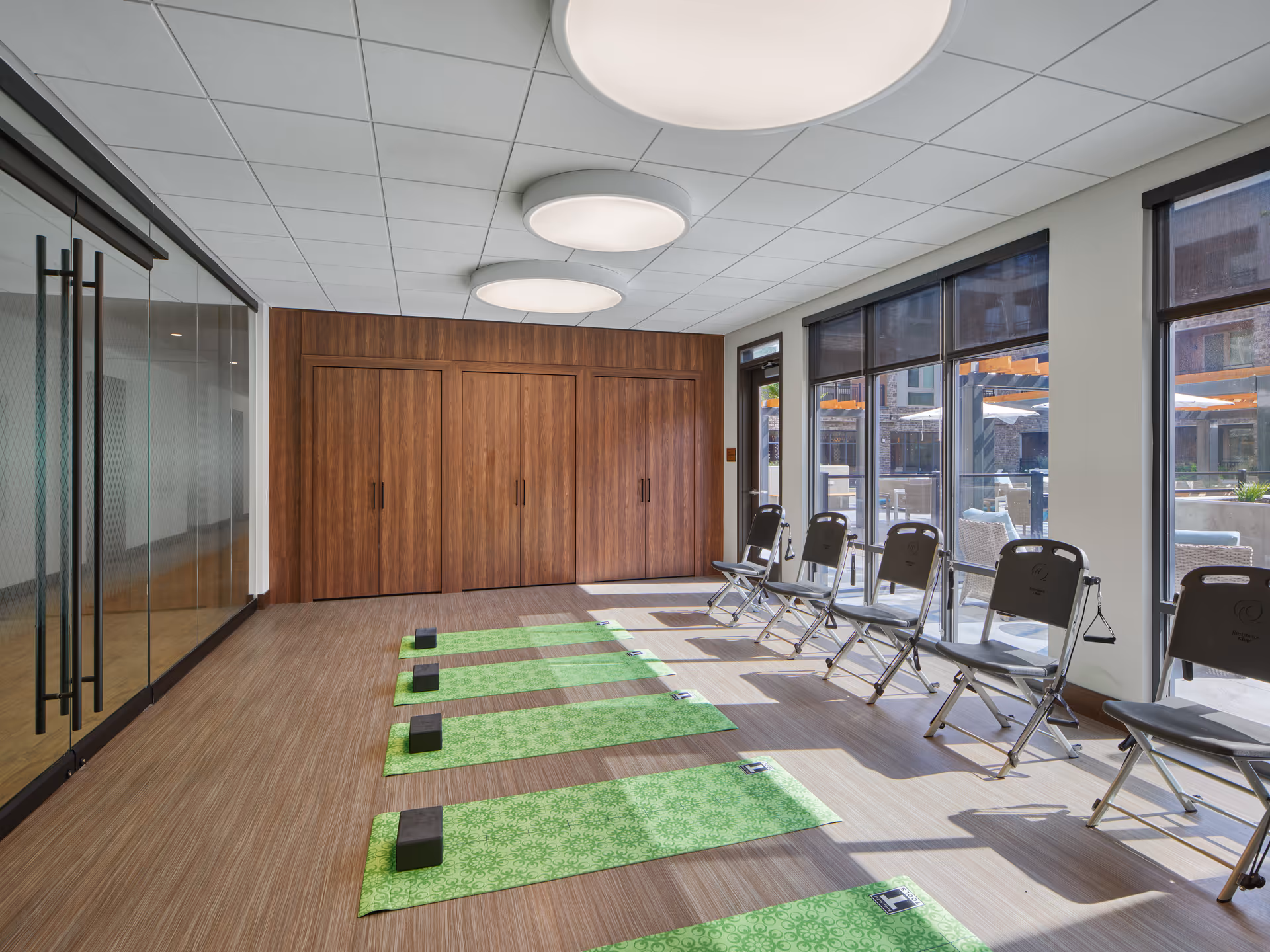 A bright exercise room with large windows letting in natural light. The room has wooden flooring and a wall with wooden paneling and closed cabinets. There are green yoga mats laid out on the floor, each with a black yoga block. Along the windows, there are several folding chairs with resistance bands attached. The ceiling has three large round light fixtures.