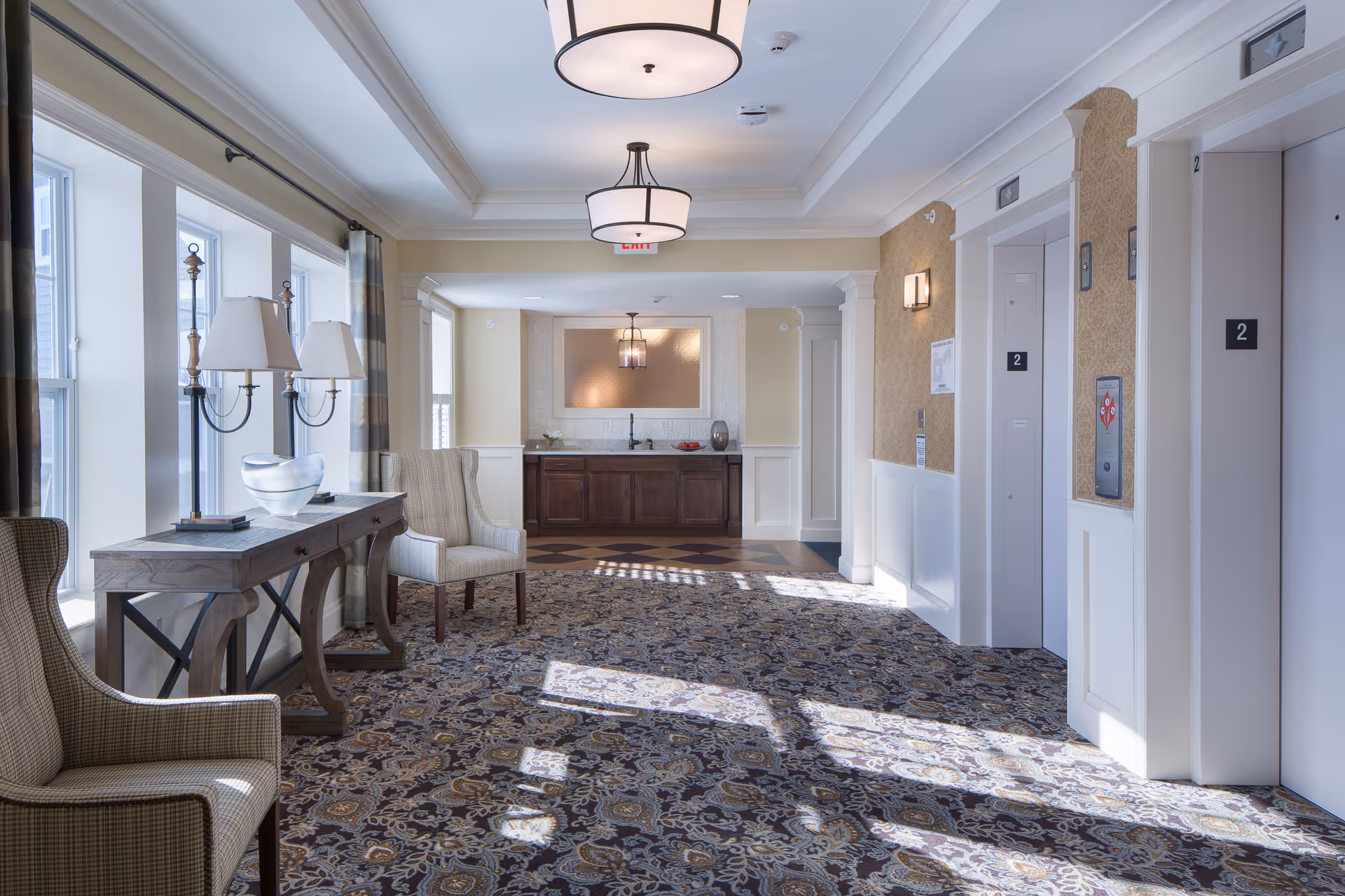 A bright and spacious hallway area in a senior living facility featuring two elevators on the right side, a patterned carpet, two upholstered armchairs, a wooden console table with two lamps and a decorative glass bowl, large windows with curtains on the left, and a small kitchenette with a sink and cabinets at the far end.