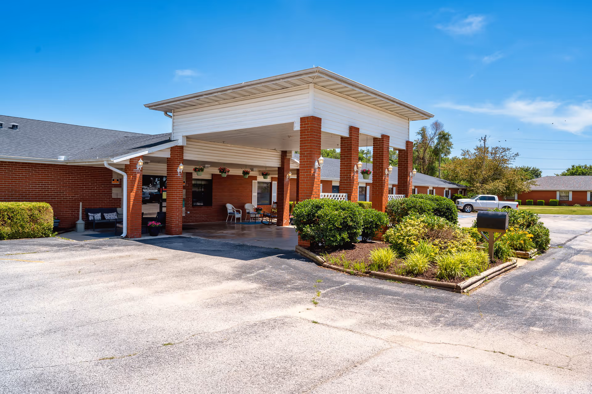 Front entrance canopy of a brick senior care facility with columns, seating, and landscaped planters under a clear blue sky.