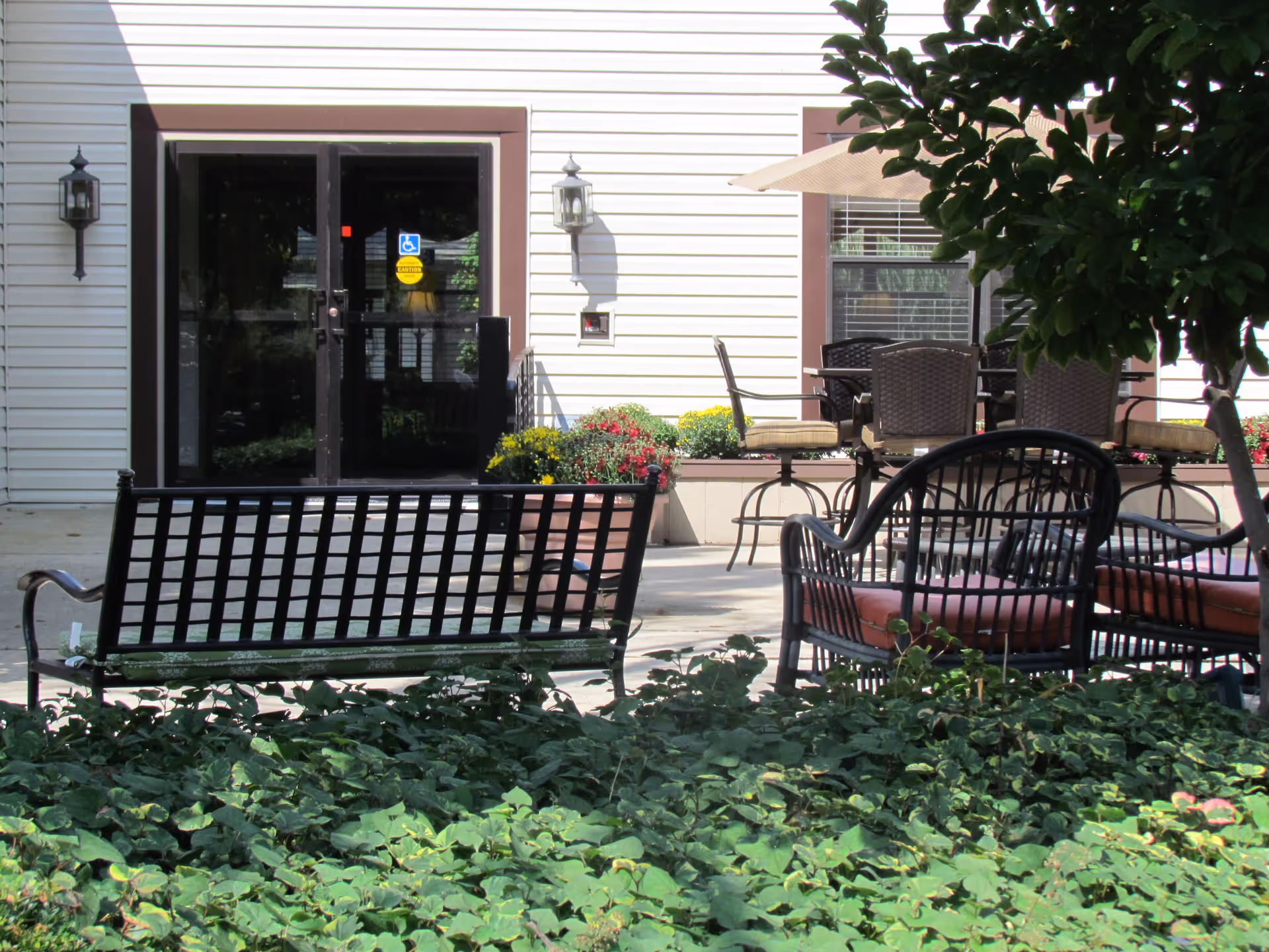 Outdoor patio area at Springfield Assisted Living featuring black metal benches, cushioned wicker chairs, a table with chairs under an umbrella, surrounded by greenery and flowers, with a building entrance in the background.