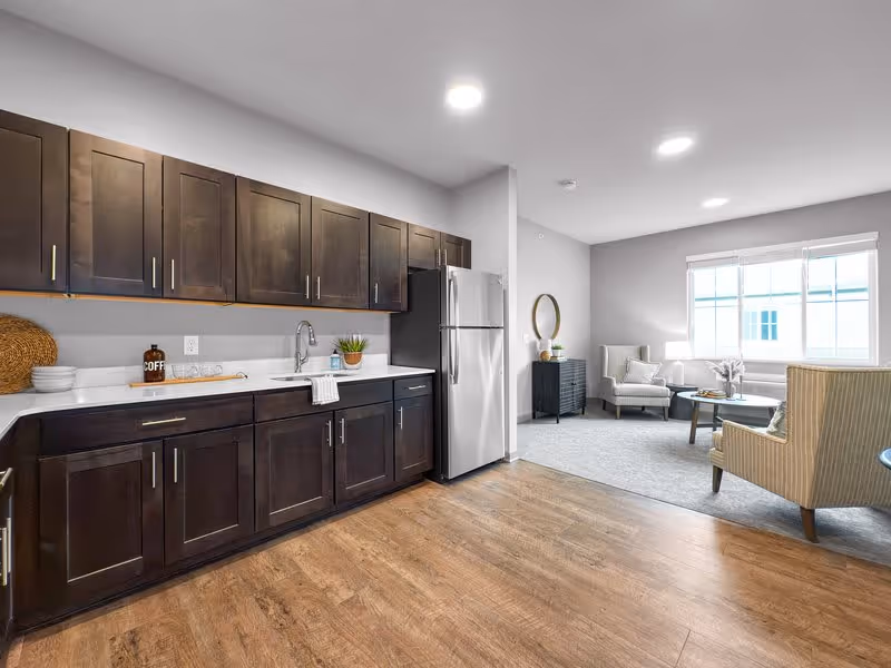 A modern kitchen area with dark wooden cabinets, a stainless steel refrigerator, and a white countertop with a sink. The kitchen opens into a living room with two armchairs, a coffee table, a side table with a lamp, and a large window letting in natural light.