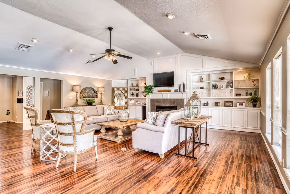 A bright and spacious living room area with wooden flooring, a ceiling fan, and recessed lighting. The room features a beige sofa, two armchairs, a wooden coffee table, and a side table with decorative lanterns. Built-in white shelves and cabinets line the far wall, with a flat-screen TV mounted above a fireplace. Large windows on the right side allow natural light to fill the space.