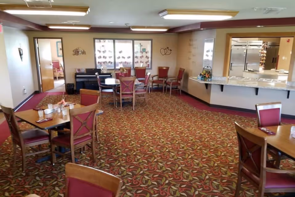 Interior view of a senior living facility dining room with several wooden tables and chairs with red cushions. The room has a patterned carpet, a piano against the far wall, and a serving counter with a kitchen visible behind it. There are windows and wall decorations, and the room is well-lit with ceiling lights.