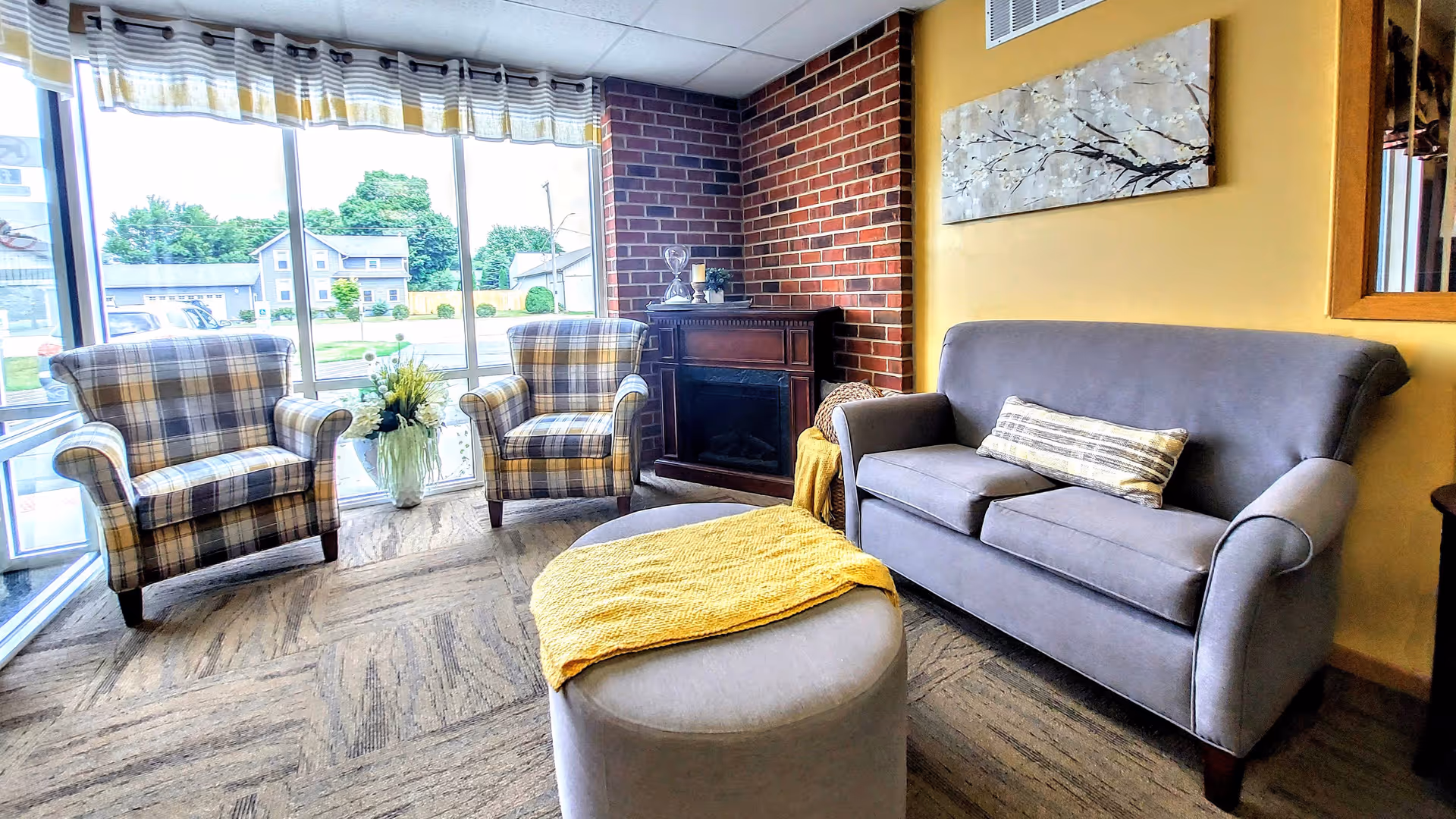 A cozy sitting area with two plaid armchairs and a gray loveseat arranged around a round gray ottoman with a yellow throw blanket. Behind the loveseat is a yellow wall with a painting of white blossoms on a tree branch. A faux fireplace with decorative items sits in the corner against a brick accent wall. Large windows with striped valances let in natural light and show a view of houses and greenery outside.