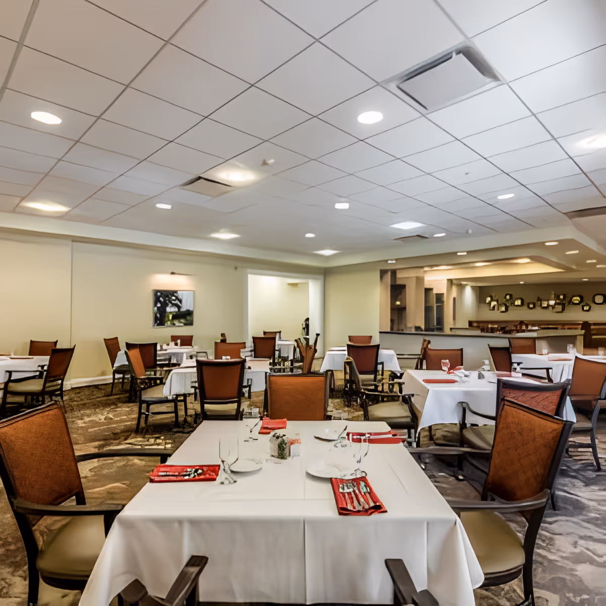 A spacious dining room with multiple tables covered in white tablecloths, each set with plates, glasses, and red napkins. The room has brown cushioned chairs, a carpeted floor, and a ceiling with recessed lighting. There is a mirror on the far wall reflecting part of the room.