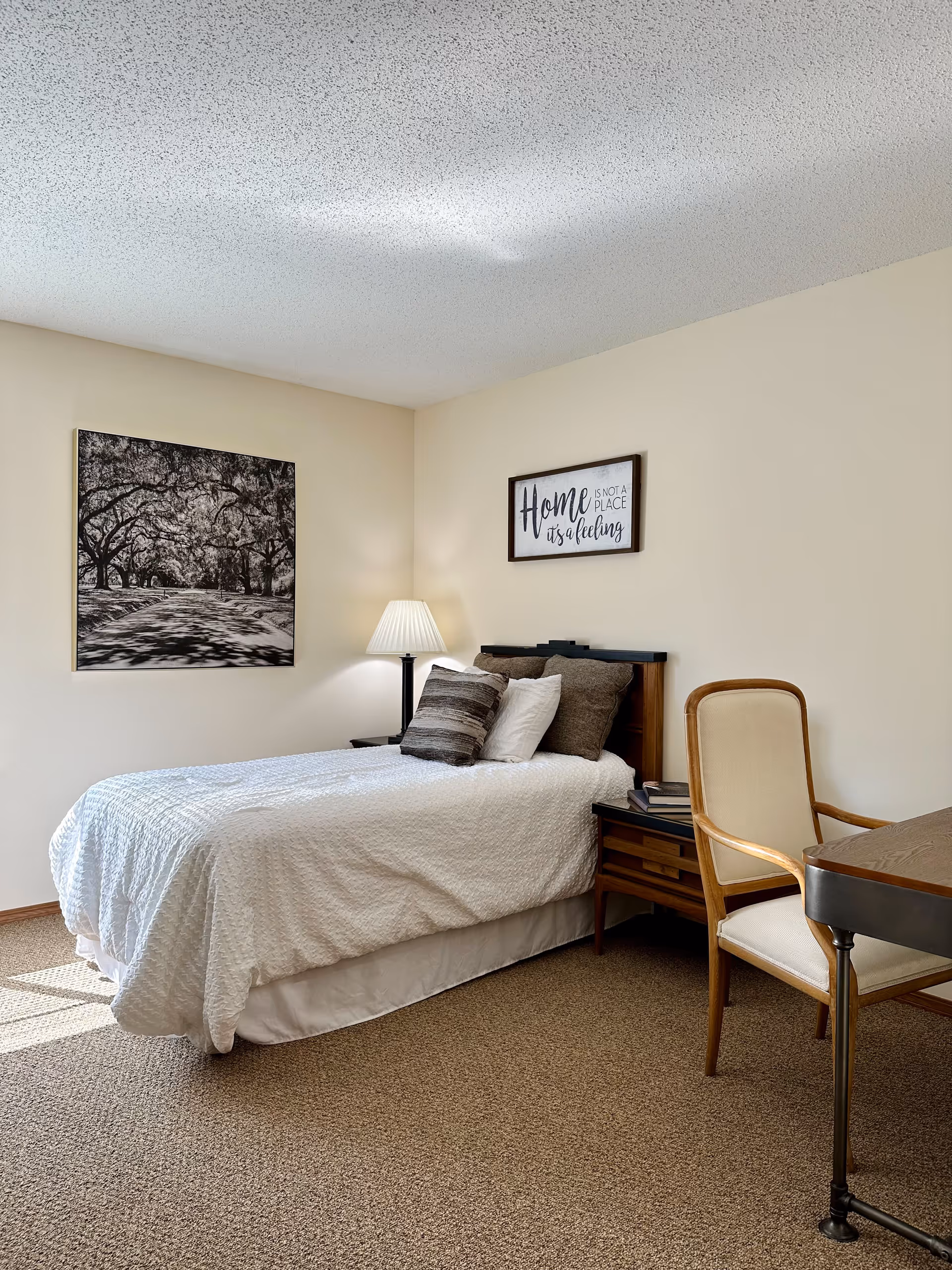 Neatly made single bed in a neutral-toned bedroom with framed wall art, a bedside lamp, nightstand and a chair at a small desk.