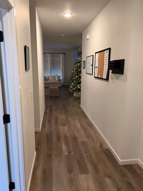 A hallway with wood flooring leading to a living room area with a beige couch and a lit Christmas tree. The walls are white with framed notices and a key holder mounted on the right side. The hallway is well-lit with ceiling lights.