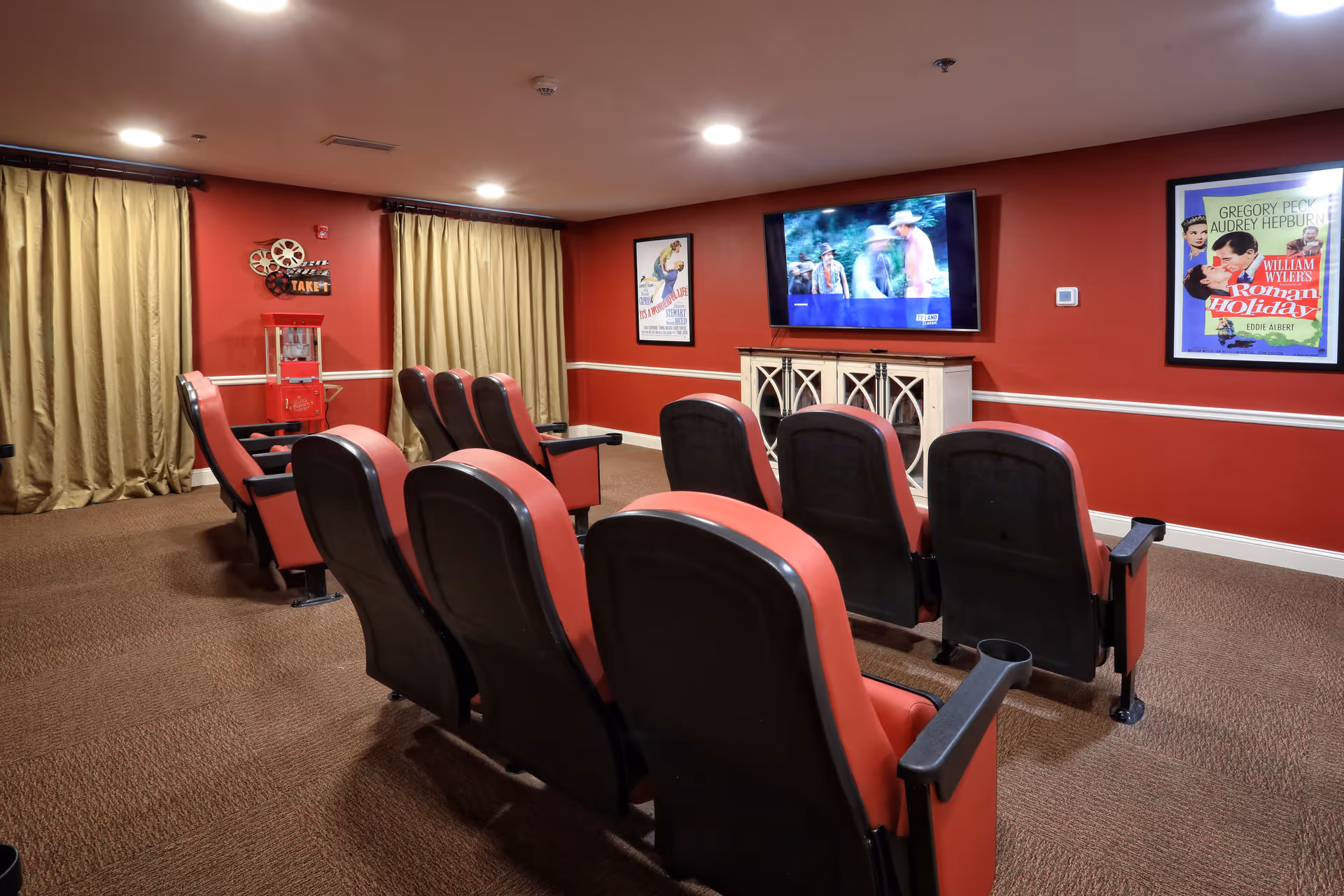 A cozy senior care facility movie room with two rows of red and black theater-style seats facing a wall-mounted flat screen TV. The room has red walls adorned with vintage movie posters, beige curtains covering the windows, and a popcorn machine in the corner.
