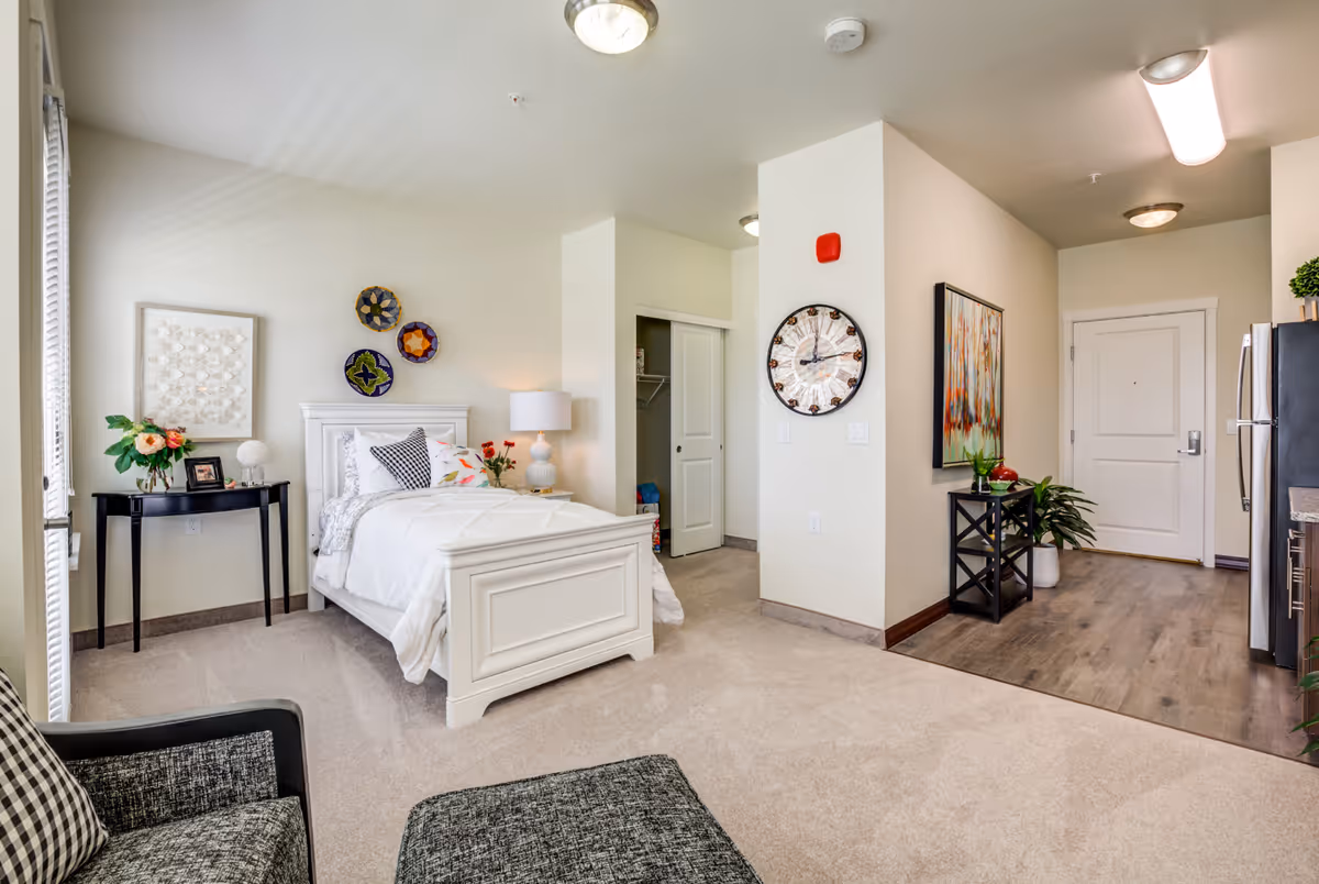 A bright and cozy senior living apartment featuring a white single bed with decorative pillows and a bedside lamp. There is a small black table with a flower vase and picture frame next to the bed. The room has beige carpet flooring and light-colored walls adorned with decorative plates and framed artwork. A large clock is mounted on a wall near the entrance door, and a small black console table with plants and decor is placed against the wall. The kitchen area with wood flooring and stainless steel appliances is visible to the right.