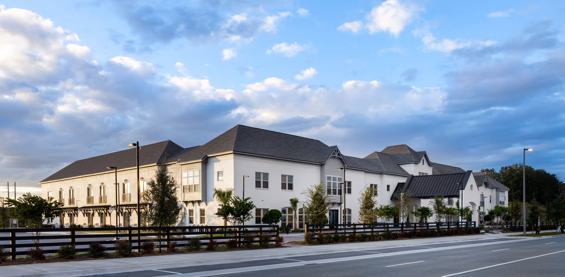 Exterior view of a large, modern senior living facility building with white walls and a dark gray roof under a partly cloudy sky. The building is surrounded by a black fence and landscaped with trees and shrubs along a paved road.
