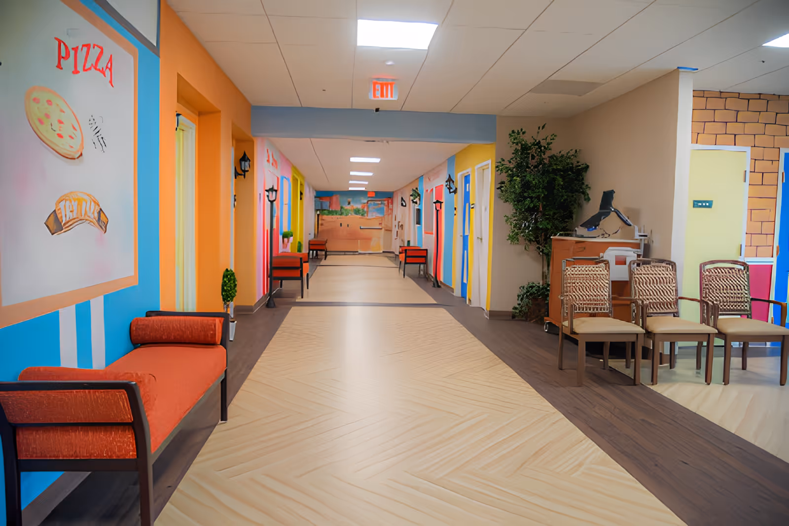 A brightly colored hallway in a senior living facility with orange, blue, yellow, and pink walls. The hallway has benches and chairs along the sides, decorative plants, and a large painting on the left wall depicting a pizza and the word 'PIZZA'. The floor has a light wood pattern with darker wood borders, and there is an exit sign on the ceiling at the end of the hallway.