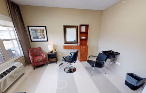 A small salon room with a styling chair in front of a mirror, a shampoo sink, and additional seating by a window.