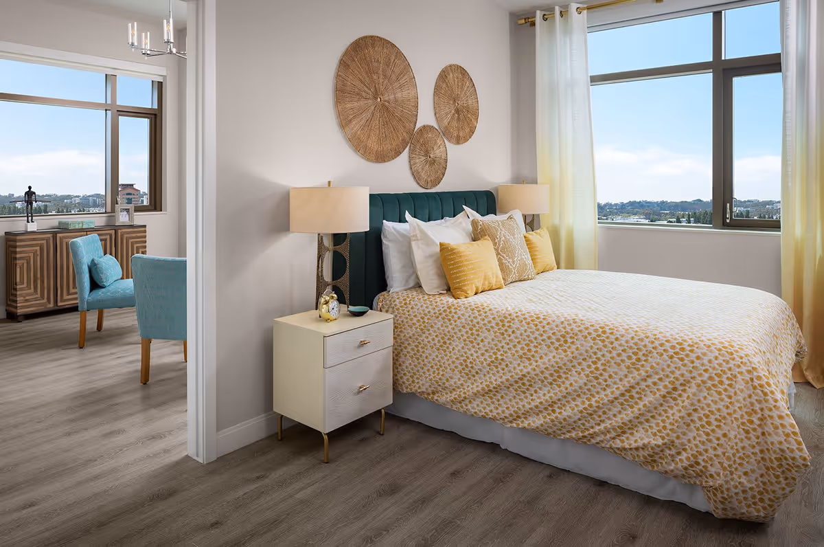 Bright, airy bedroom with a bed covered in yellow-patterned bedding, a nightstand and lamps, large windows with a view, and an adjacent seating area visible through a doorway.