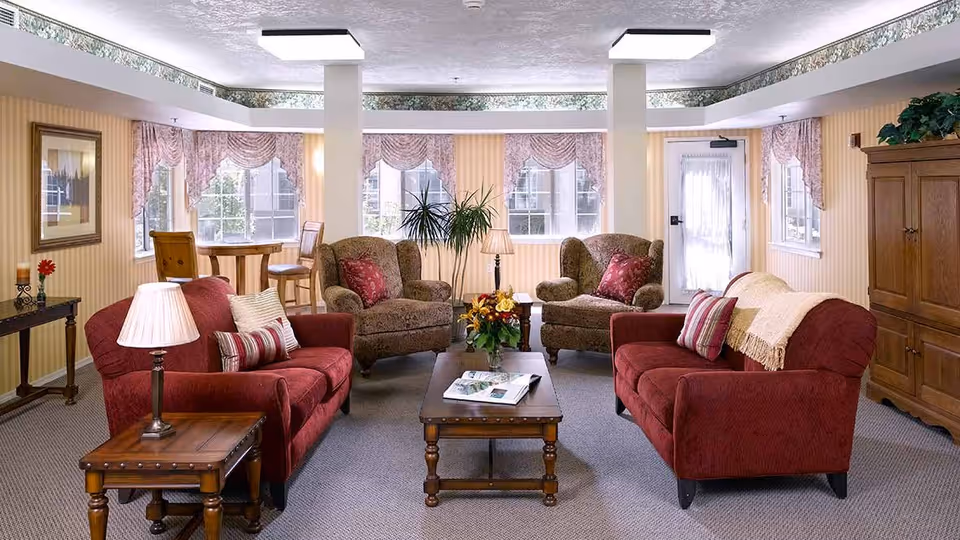 A cozy living room with two red sofas and two patterned armchairs arranged around a wooden coffee table with a flower arrangement and magazines. The room has large windows with pink valances, a wooden side table with a lamp, a wooden cabinet, and a small round table with chairs near the windows. The walls are decorated with striped wallpaper and a framed picture.