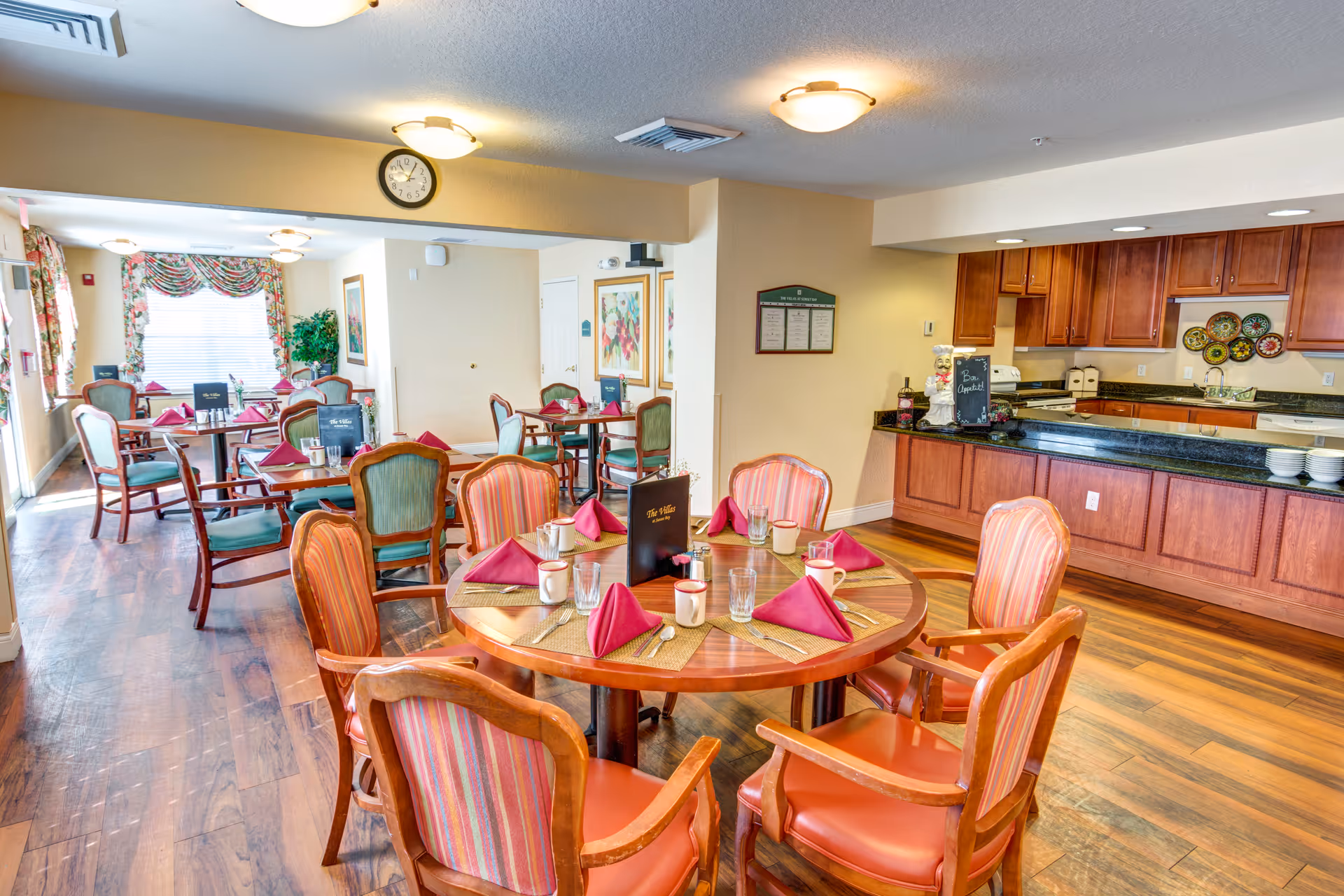 Bright dining room with round tables set with napkins and cups and a serving counter/kitchen area in the background.
