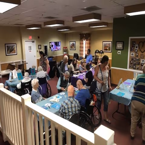 A group of elderly residents and caregivers gathered in a well-lit common area with tables covered in blue and purple tablecloths. Some residents are seated in wheelchairs while others sit in chairs, engaging in activities or conversations. The room has beige walls with framed pictures and a bulletin board, and a television is mounted on the far wall.