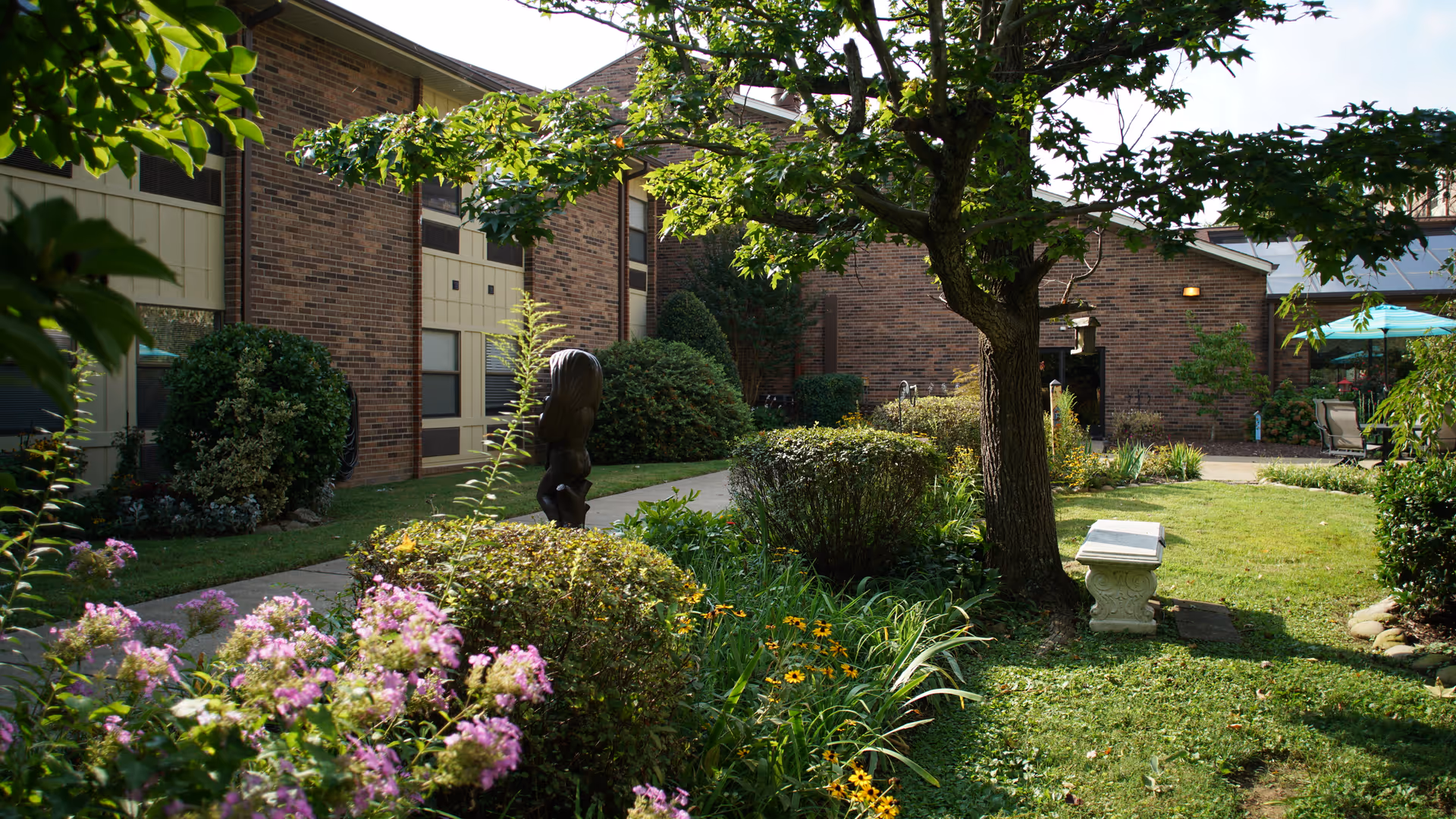 Sunlit courtyard garden with a tree, flowering plants, a stone bench and a brick retirement community building in the background.