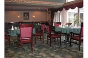 Dining room with multiple tables covered with green tablecloths, set with white napkins and glassware. The room has wooden chairs with red cushions, patterned carpet, and a large window with maroon curtains allowing natural light in.