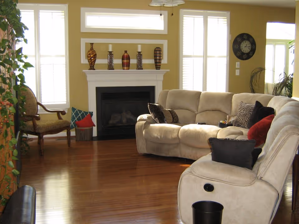 A cozy living room with a beige sectional sofa adorned with various throw pillows in black, red, and patterned designs. The room features a white fireplace mantel decorated with vases and candle holders. Large windows with white blinds allow natural light to fill the space. A wooden chair with green upholstery is placed near the window, and a round wall clock is visible on the wall. The floor is polished hardwood, and there are some green plants adding a touch of nature to the room.