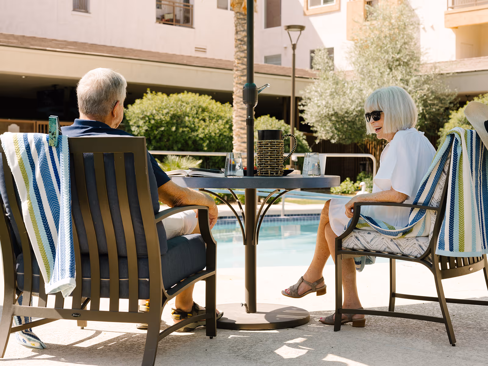 An elderly man and woman sitting outdoors at a round table by a swimming pool. The man is wearing a navy blue shirt and white shorts, and the woman is wearing a white dress and sunglasses. Both chairs have striped towels draped over the back. There are glasses and a pitcher on the table, and the background shows greenery and a multi-story building.