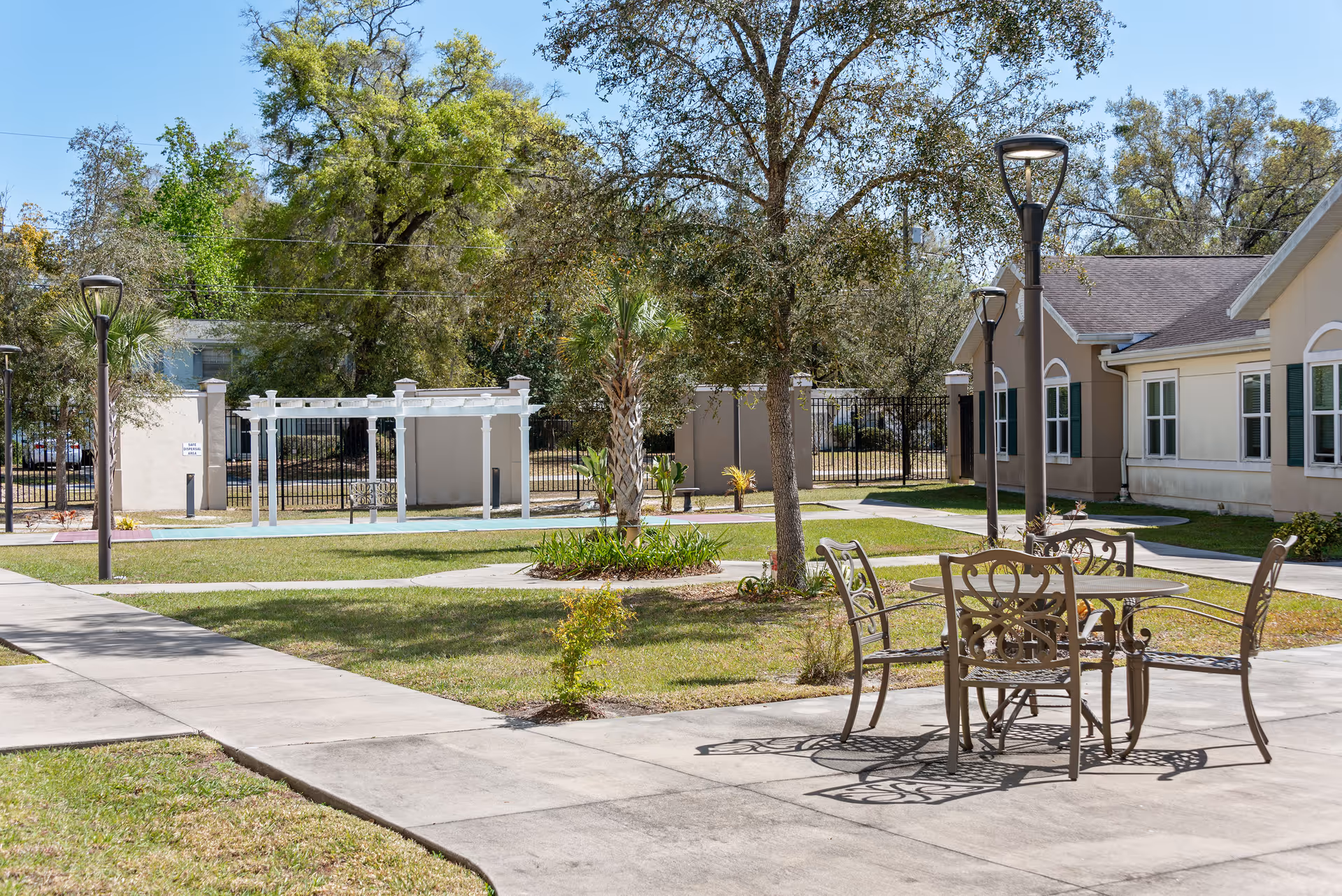 Outdoor courtyard with a metal table and chairs, paved walkways, lamp posts, trees, and surrounding single-story buildings.