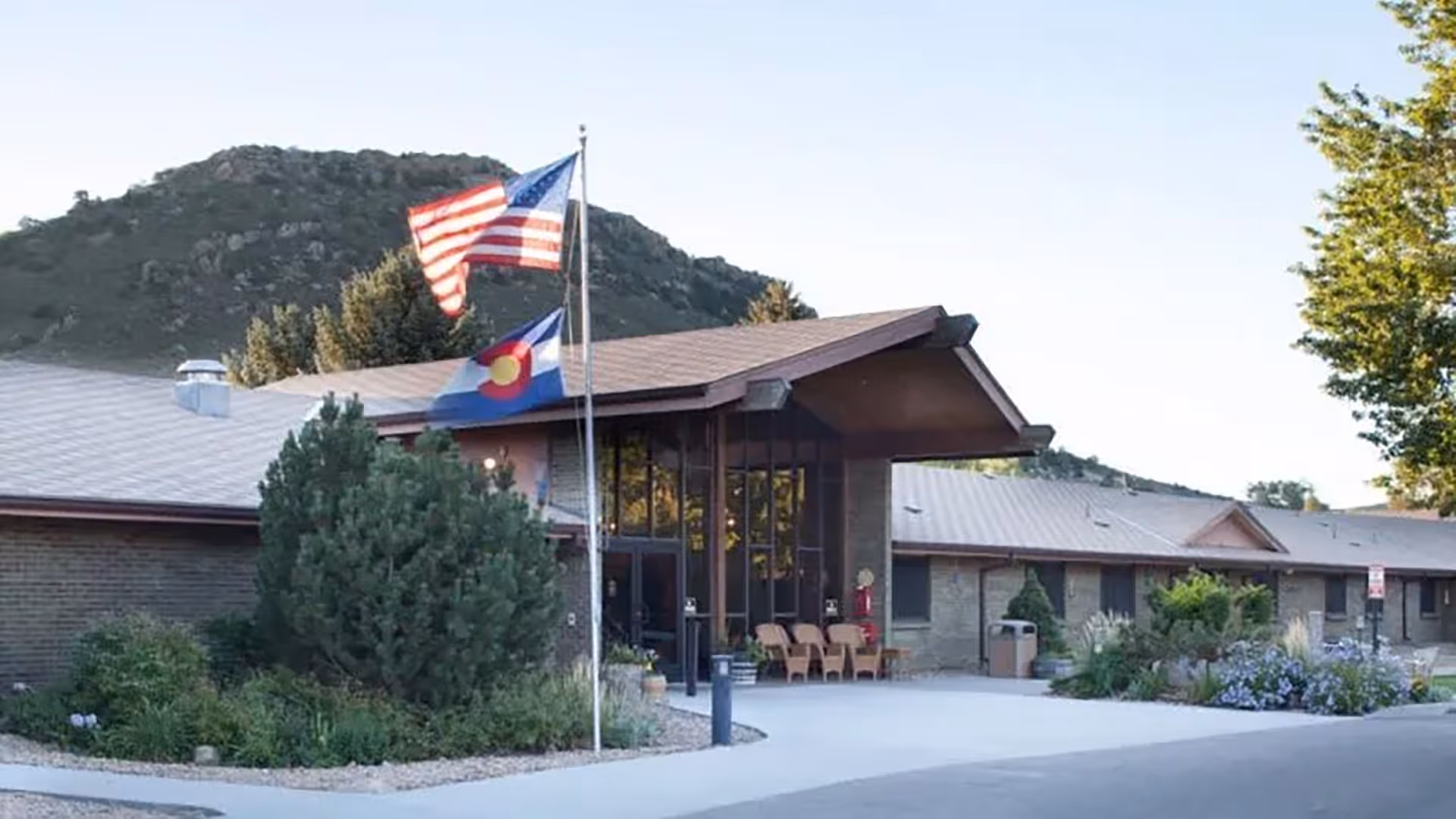 Exterior view of Prestige Care Center of Morrison, a single-story building with a sloped roof, surrounded by greenery and mountains in the background. Two flagpoles display the American flag and the Colorado state flag near the entrance.
